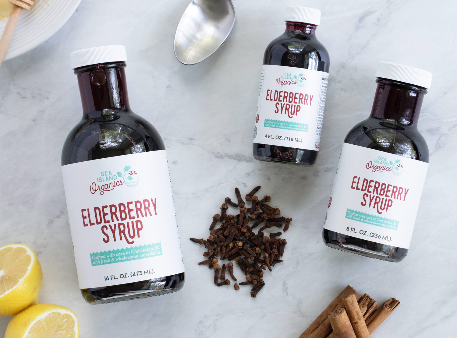 Three sizes of elderberry syrup and various ingredients on a marble countertop.
